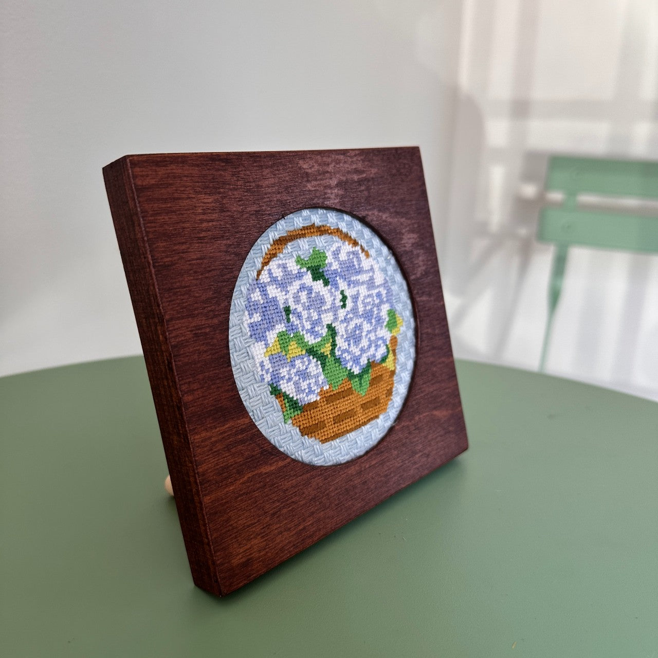 Wooden box with a cross-stitched design of a basket of flowers on a green surface.