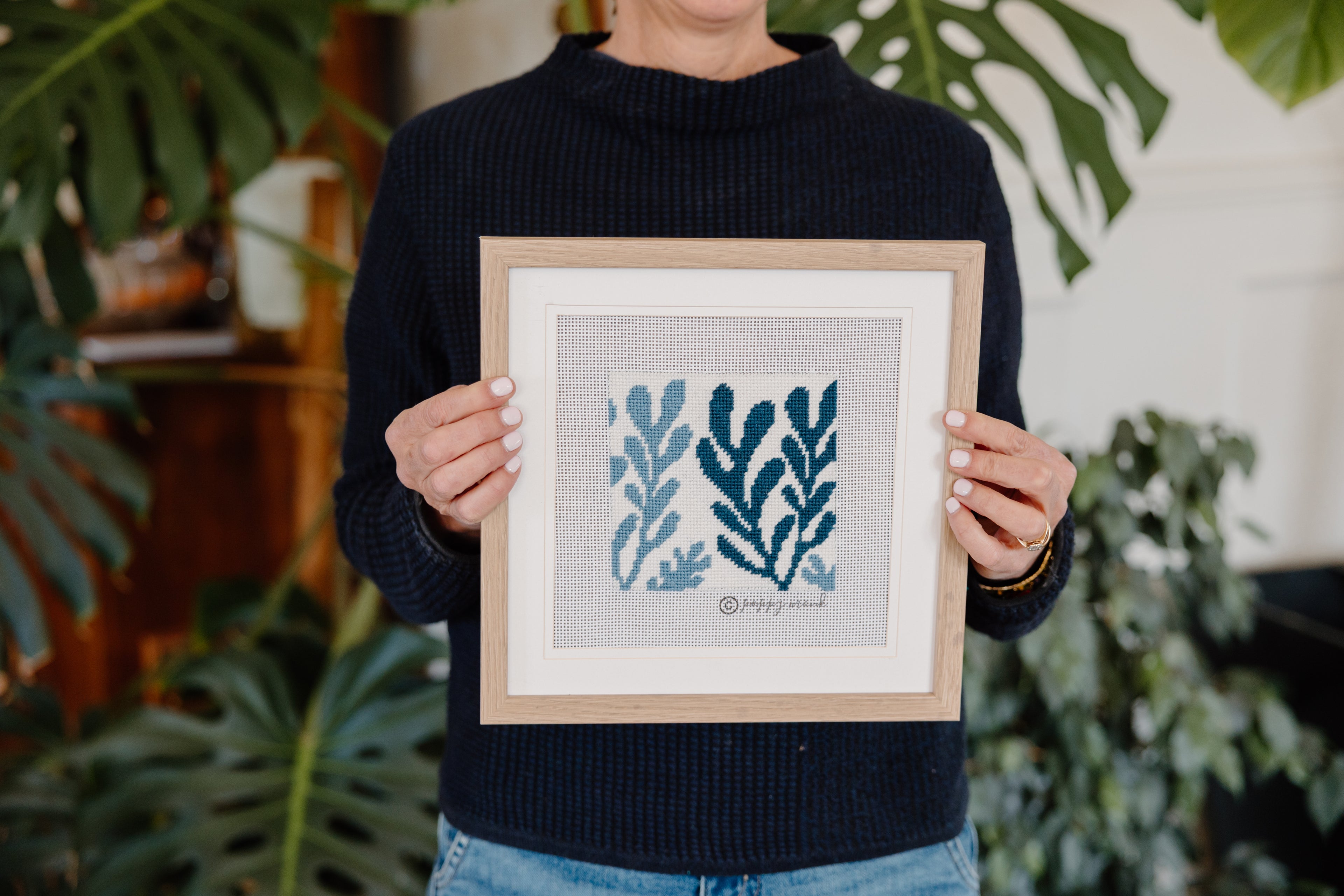 Person holding a framed needlepoint artwork with blue leaf designs against a plant-filled background