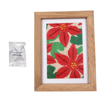 Framed needlepoint poinsettia design with a packet of hanging hardware on a white background