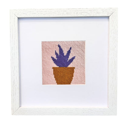Framed needlepoint artwork of a purple plant in a brown pot on a white background