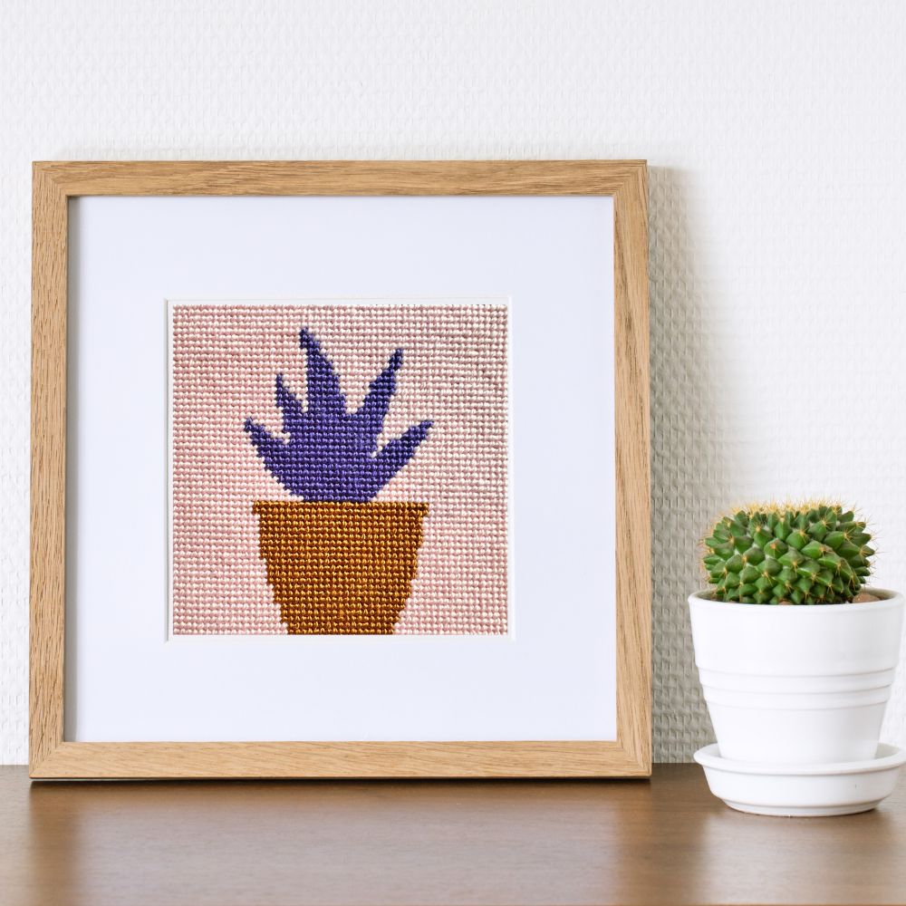 Framed needlepoint art of a potted plant on a table with a real cactus.