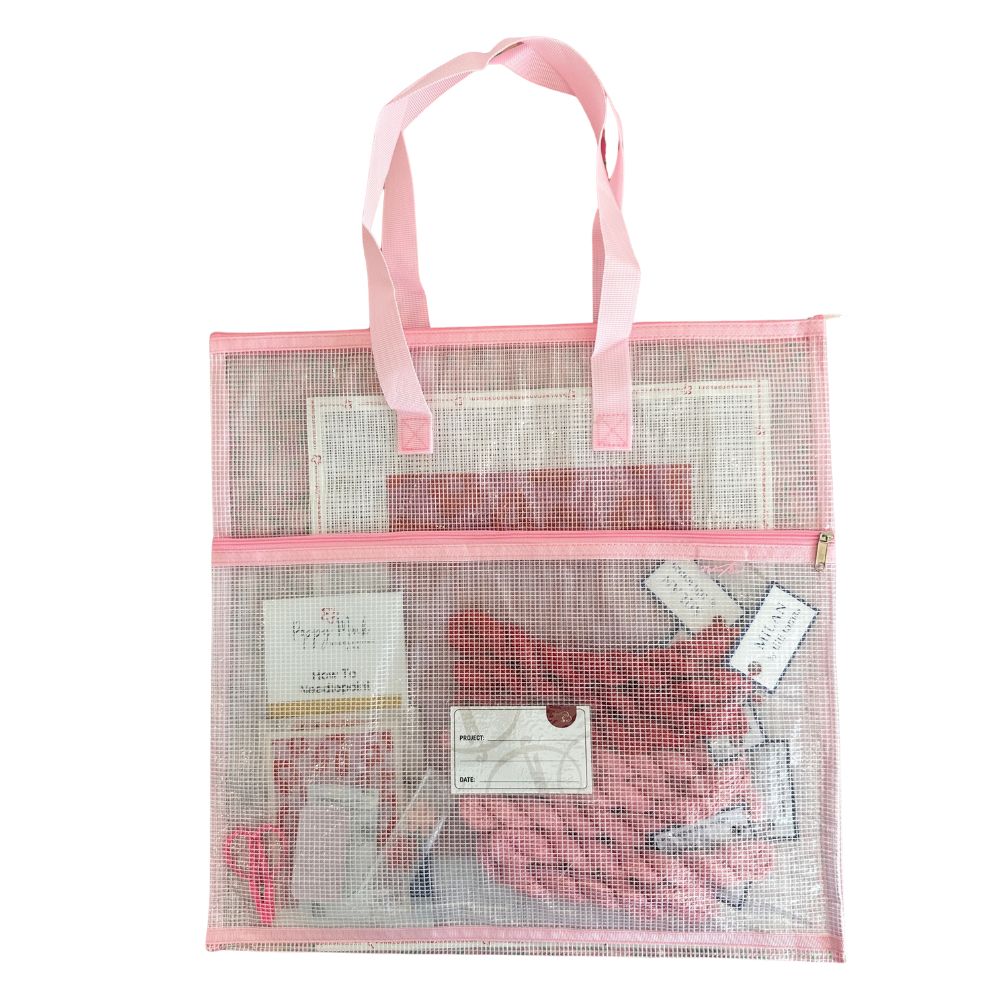 Mesh tote bag with pink handles filled with needlepoint items on a white background