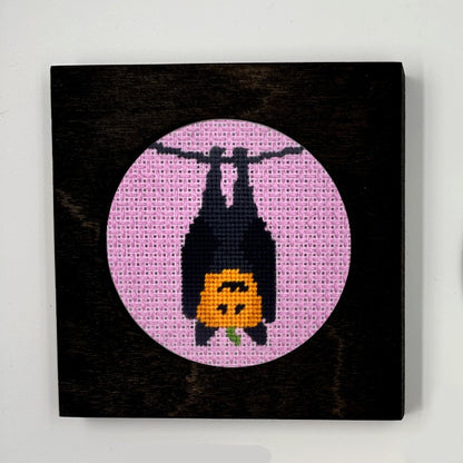 A Halloween needlepoint design of a pumpkin in a bat costume, displayed in a frame