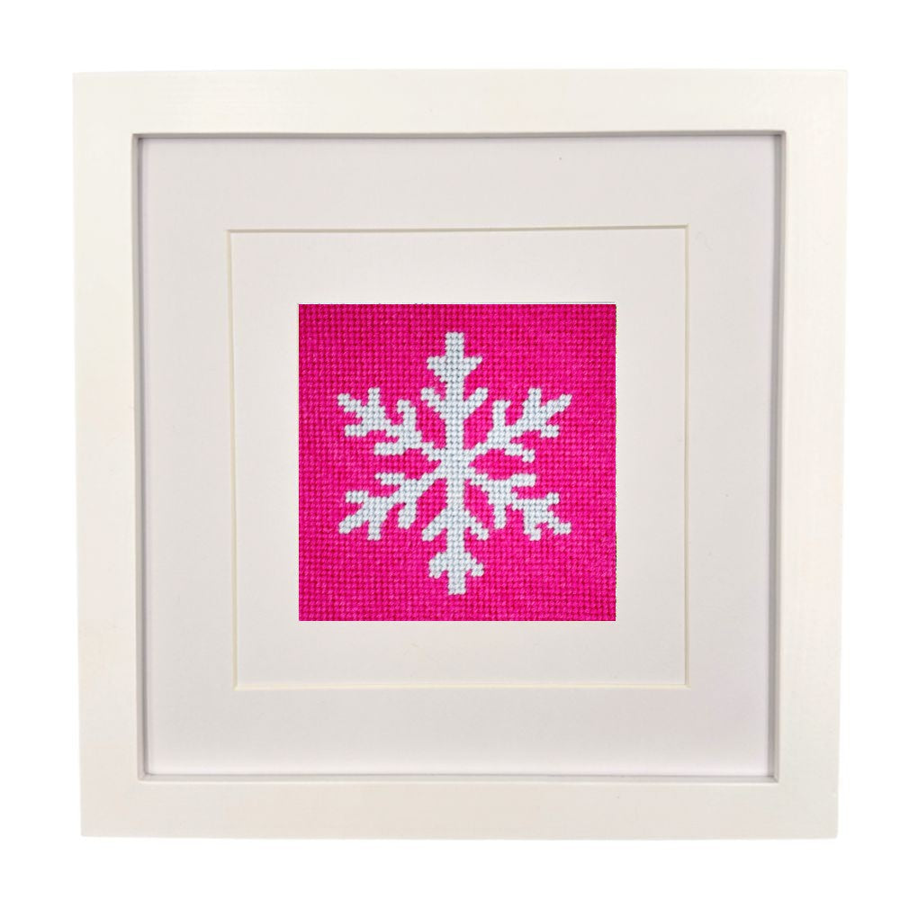 A square framed art piece featuring a white needlepoint snowflake on a bright pink fabric background, set within a white mat and a white wooden frame.