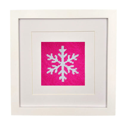 A square framed art piece featuring a white needlepoint snowflake on a bright pink fabric background, set within a white mat and a white wooden frame.