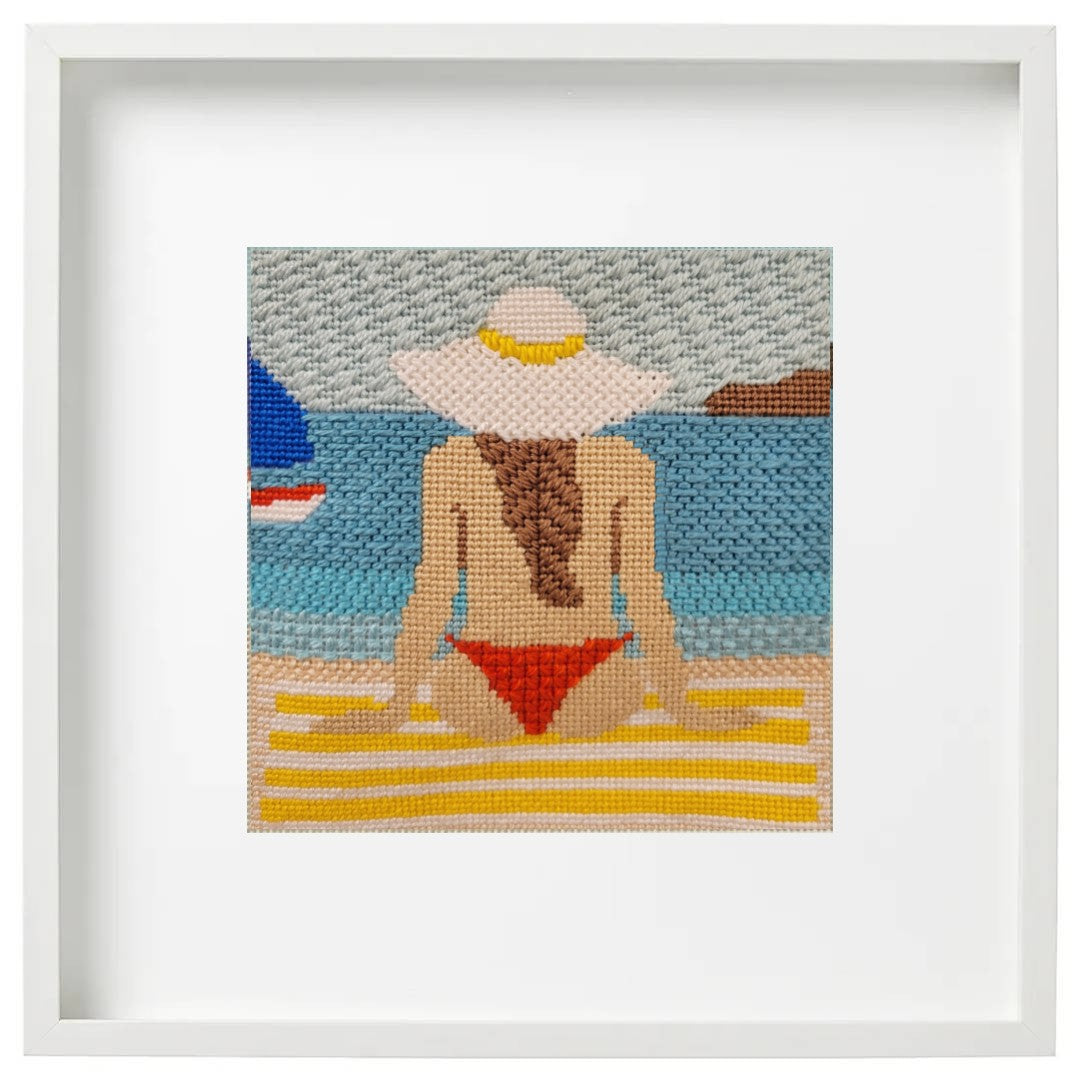 Framed needlepoint art of a woman in a red bikini and white sun hat on a beach.