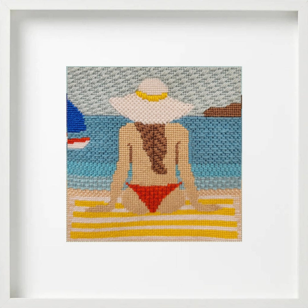 Framed needlepoint art of a woman in a red bikini and white sun hat on a beach.