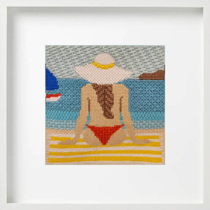Framed needlepoint art of a woman in a red bikini and white sun hat on a beach.